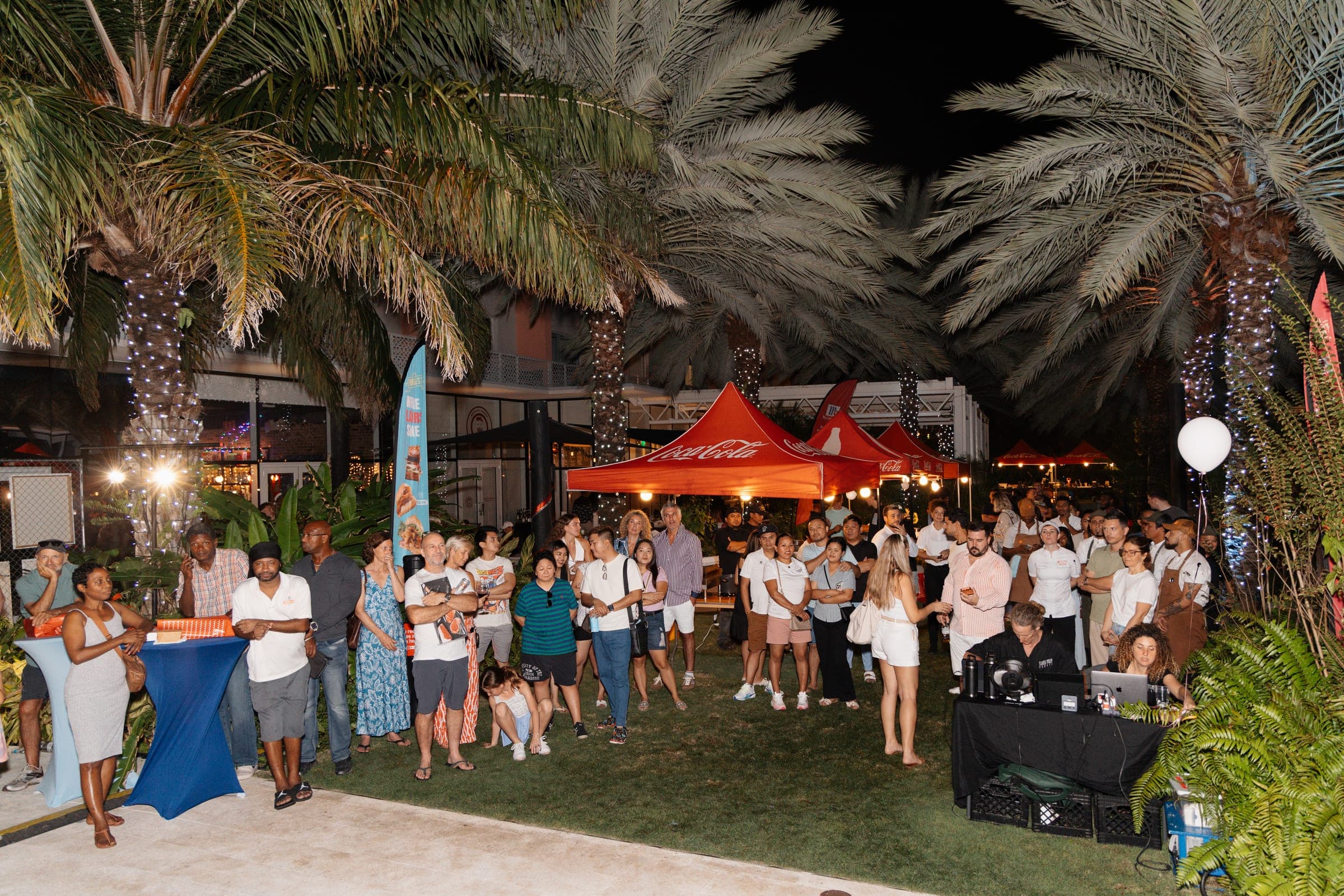 Night crowd gathered on a palm-lined lawn in front of red Coca-Cola tents at the Cayman Burger Fest Grand Finale