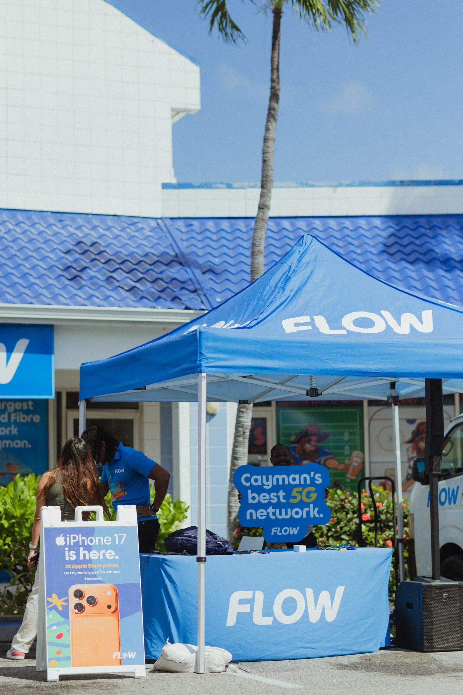 A blue Flow pop-up tent and branded table outside a Flow store, with an iPhone 17 sign and a Cayman’s best 5G network placard