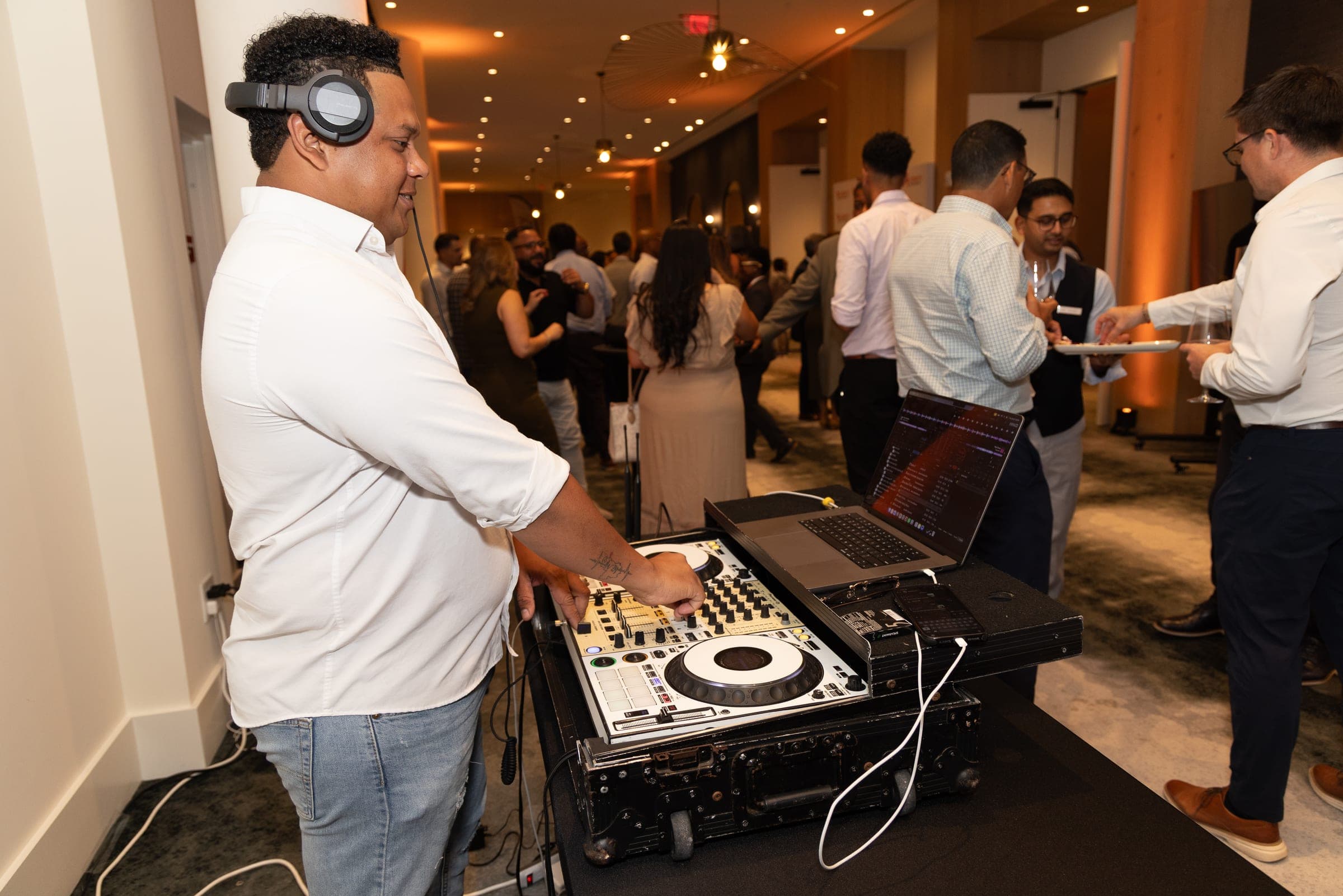 A DJ plays in the foreground while a crowd of guests mingles in the corridor behind