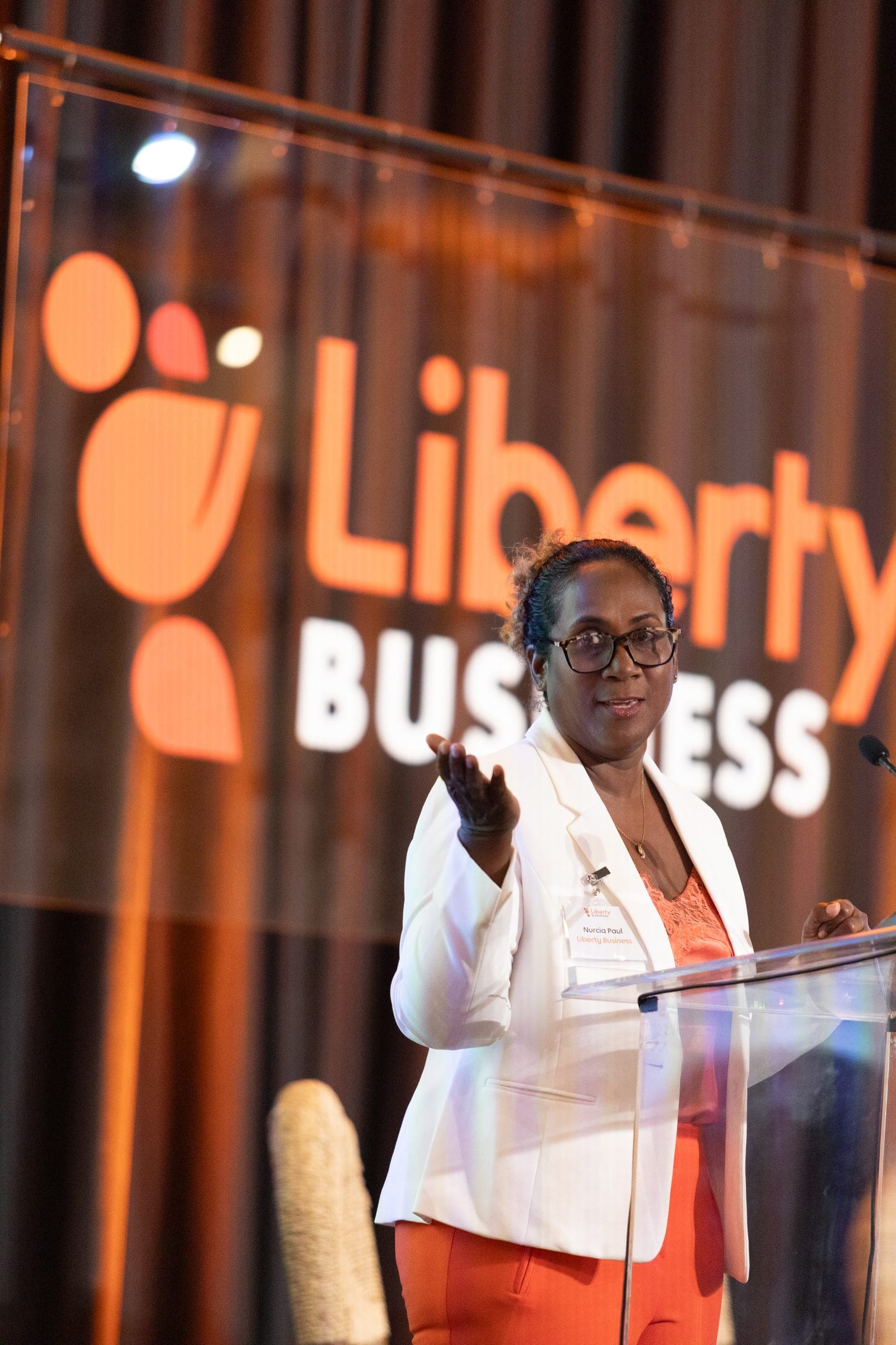 A speaker in a white blazer gestures from the podium beneath an illuminated Liberty Business logo