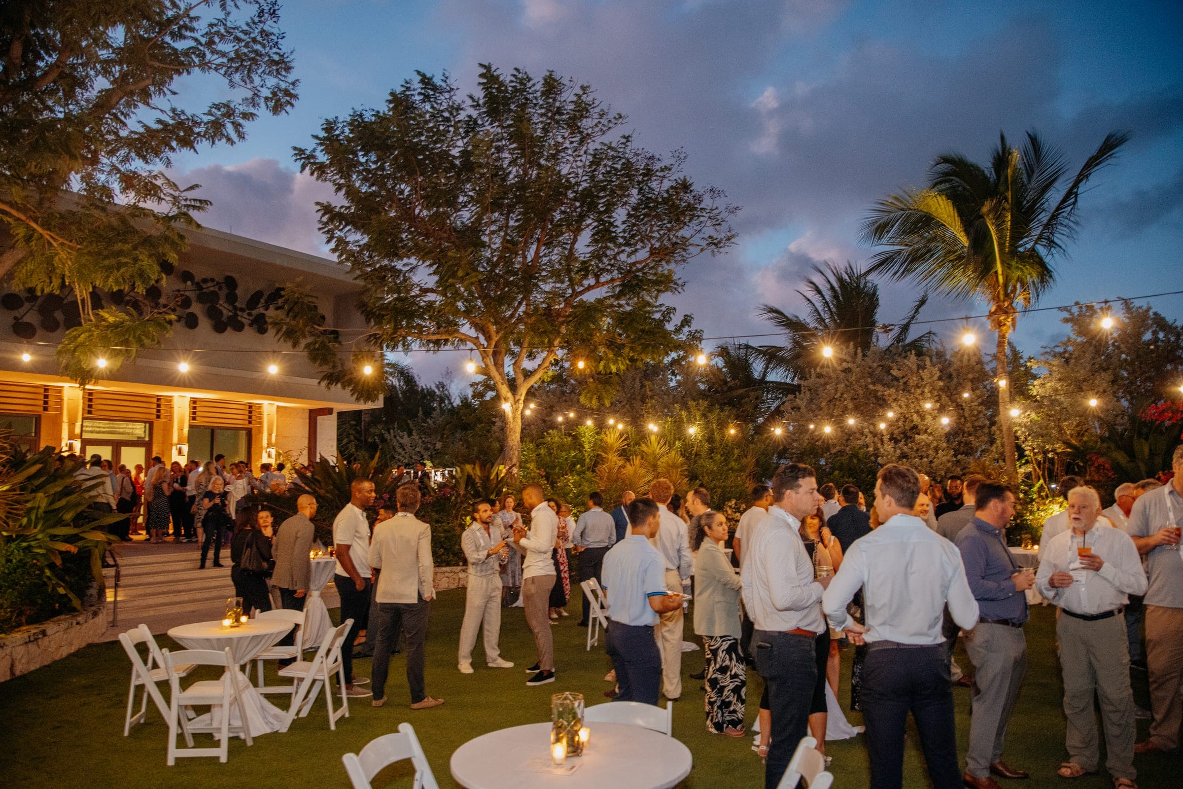Guests mingle on a lawn under string lights and palm trees at dusk during the STEP Cayman Conference welcome reception