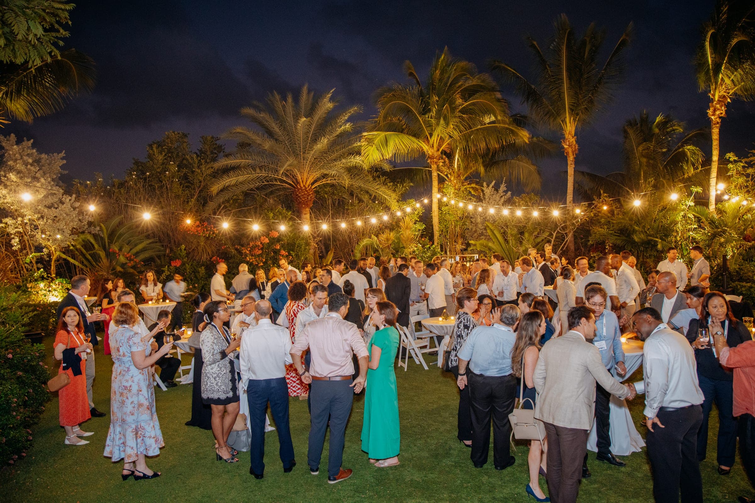 A large crowd of conference delegates networking at night in a garden lit by festoon lights