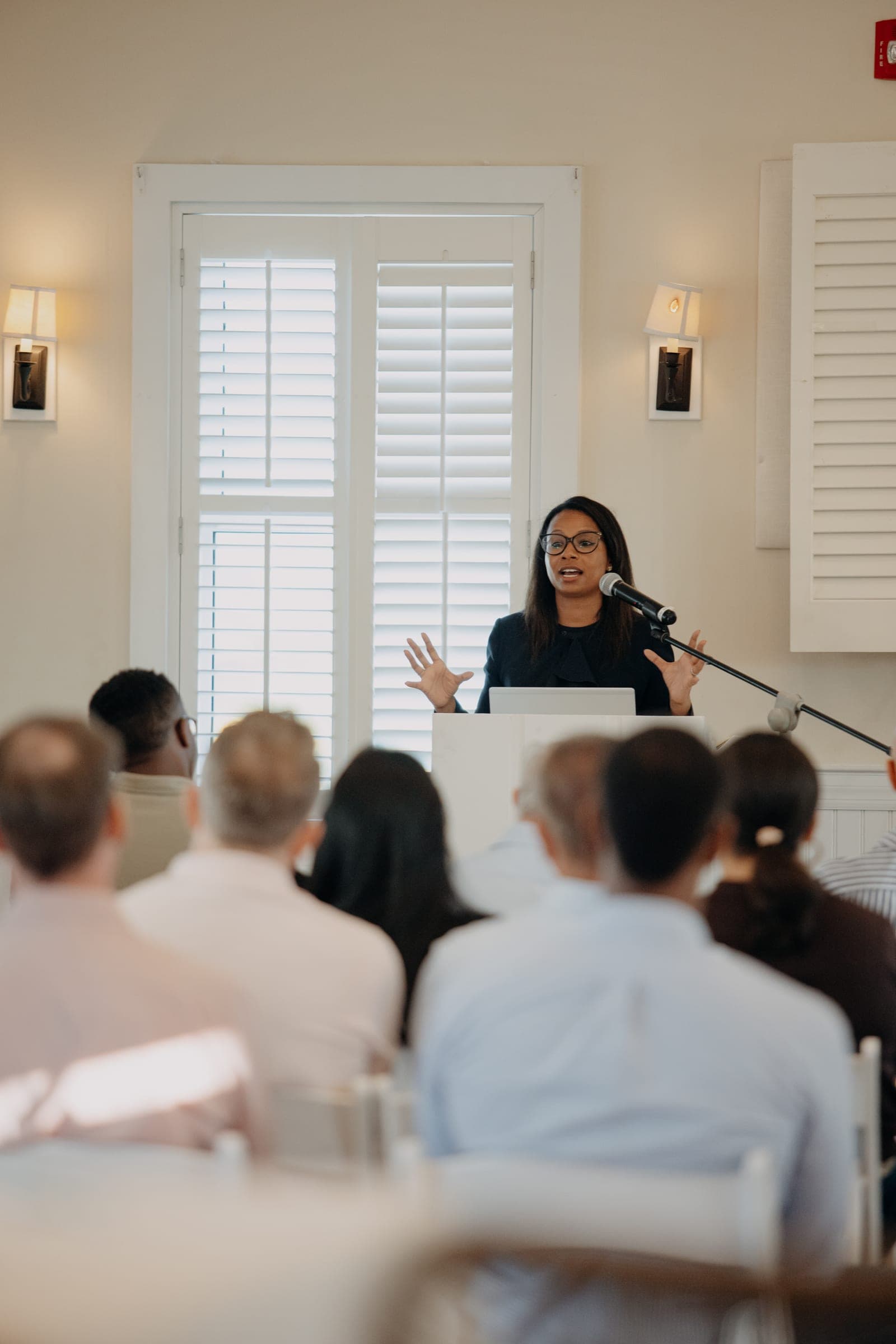 A speaker addresses a seated audience from a lectern in a bright room with white shutters