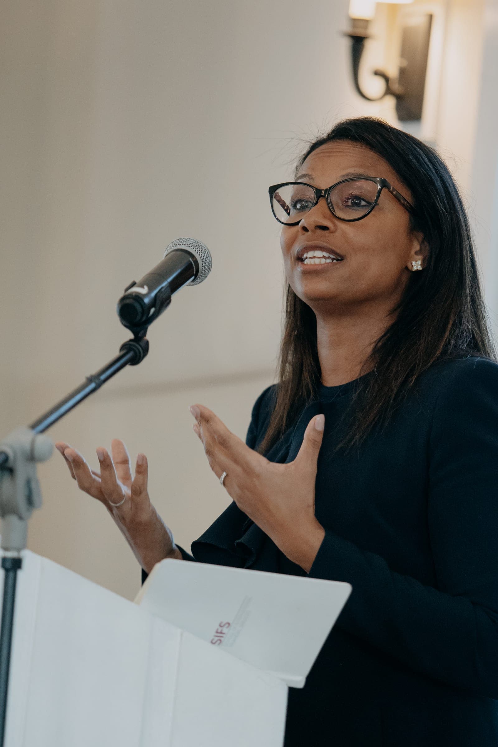 A speaker in glasses gestures while talking into a microphone at a lectern